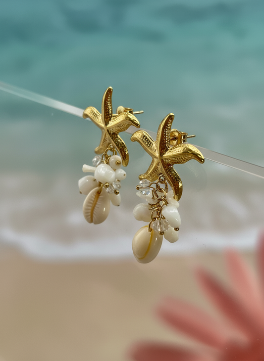Gold starfish dangle earrings with white seashell and pearl charms on beach background