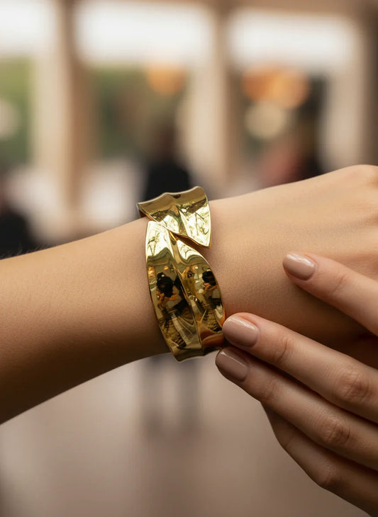 A gold-plated, wide cuff bracelet with a modernist, sculptural design featuring two overlapping, curved bands that resemble leaves or waves. The bracelet has a shiny, sculptural gold tone.